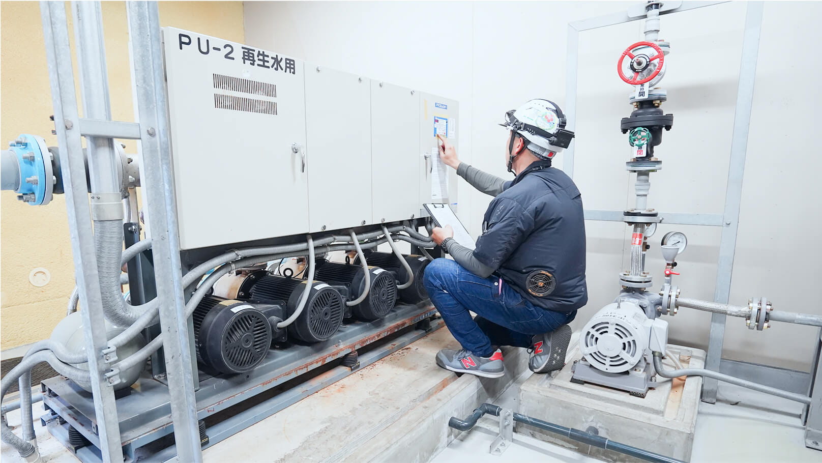 作業服とヘルメットを着用した作業員が、再生水用ポンプ装置の操作盤を点検している様子